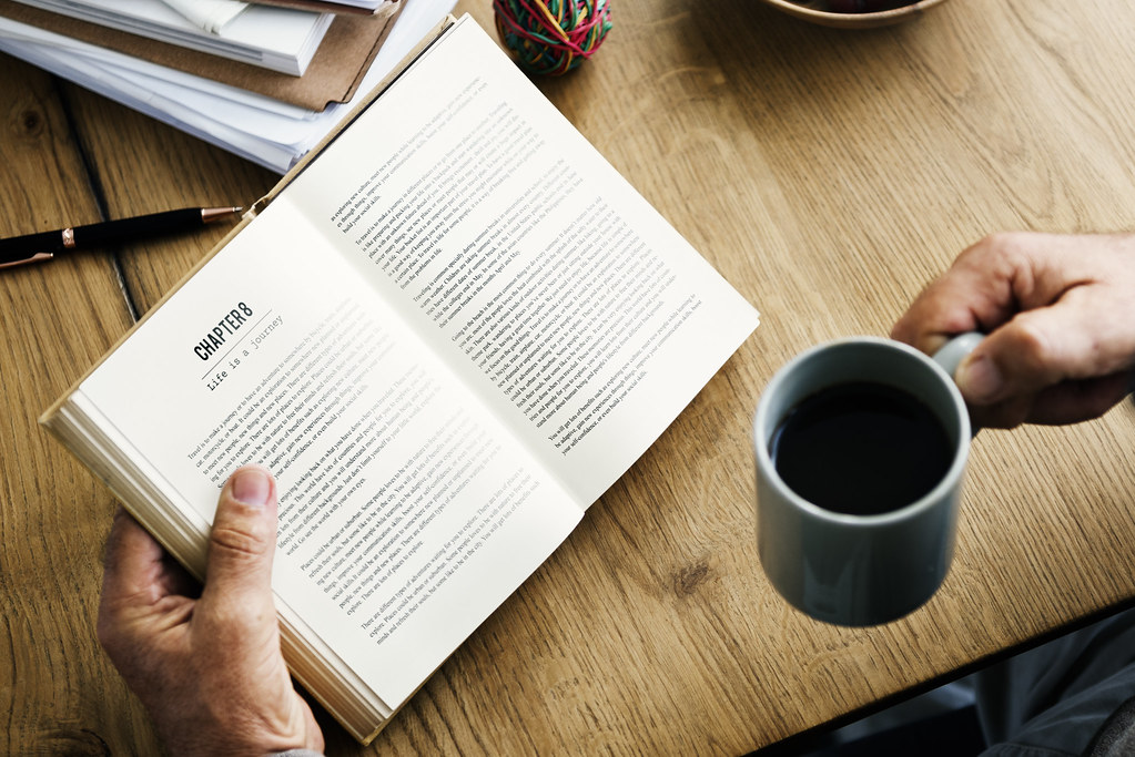 Hands holding an open book at a chapter page beside a mug of coffee on a wooden table